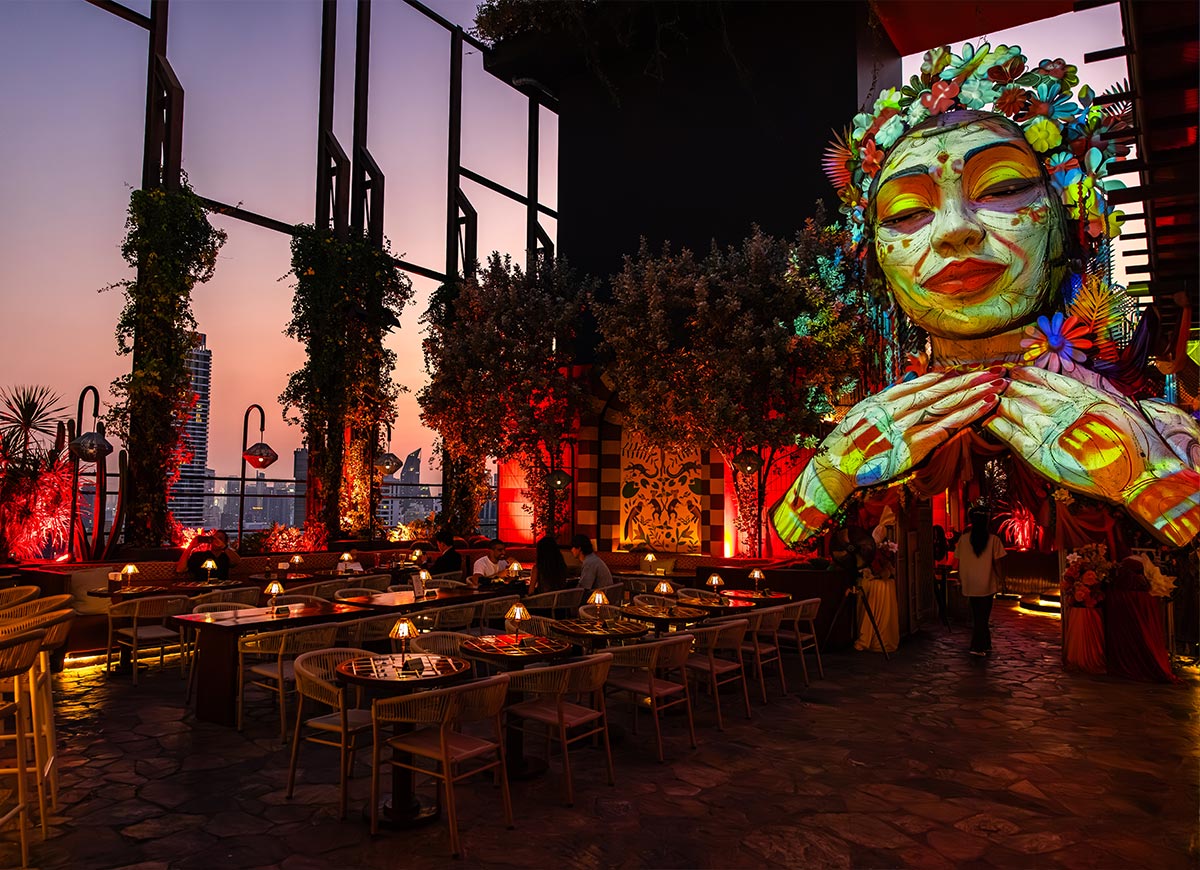 Stylish rooftop restaurant at sunset with warm red lighting and lush greenery surrounding dining tables. A large colorful sculpture of a serene face with floral details overlooks the space, creating a vibrant and artistic atmosphere.