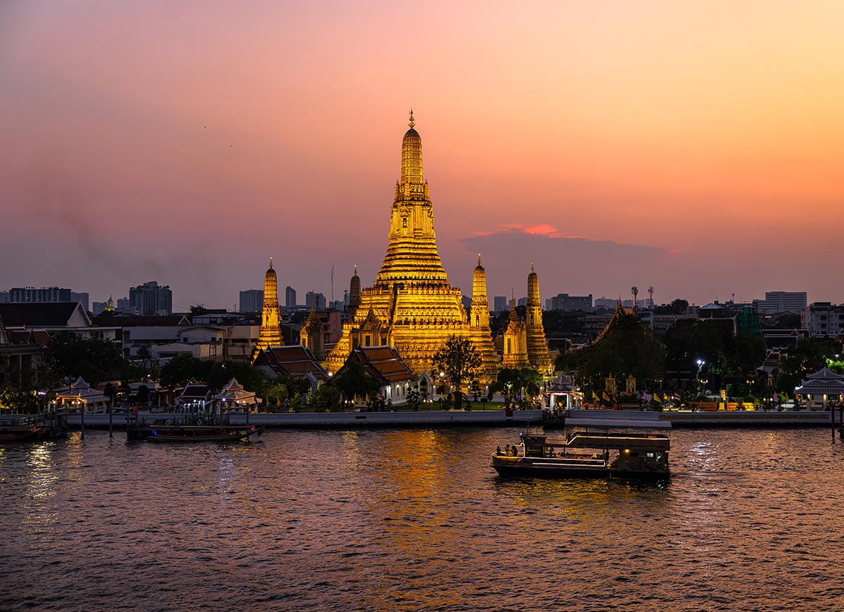 Wat Arun temple glowing with golden lights along the Chao Phraya River at sunset in Bangkok. A small boat cruises across the water in front of the illuminated temple while the sky fades from orange to soft pink at dusk.