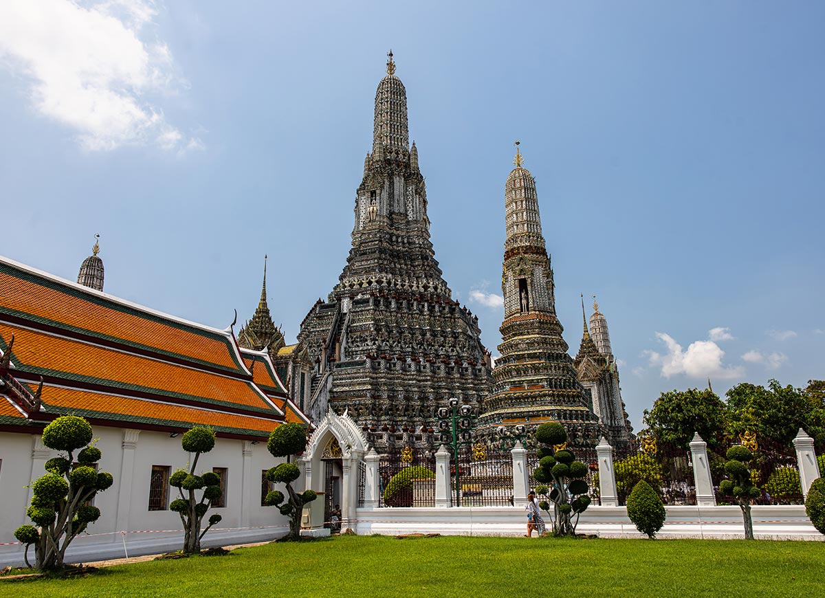 Intricately decorated temple towers rise behind a white wall and manicured garden, with detailed mosaic patterns covering the tall spires. A person walks along the path in front of the complex under a bright blue sky.