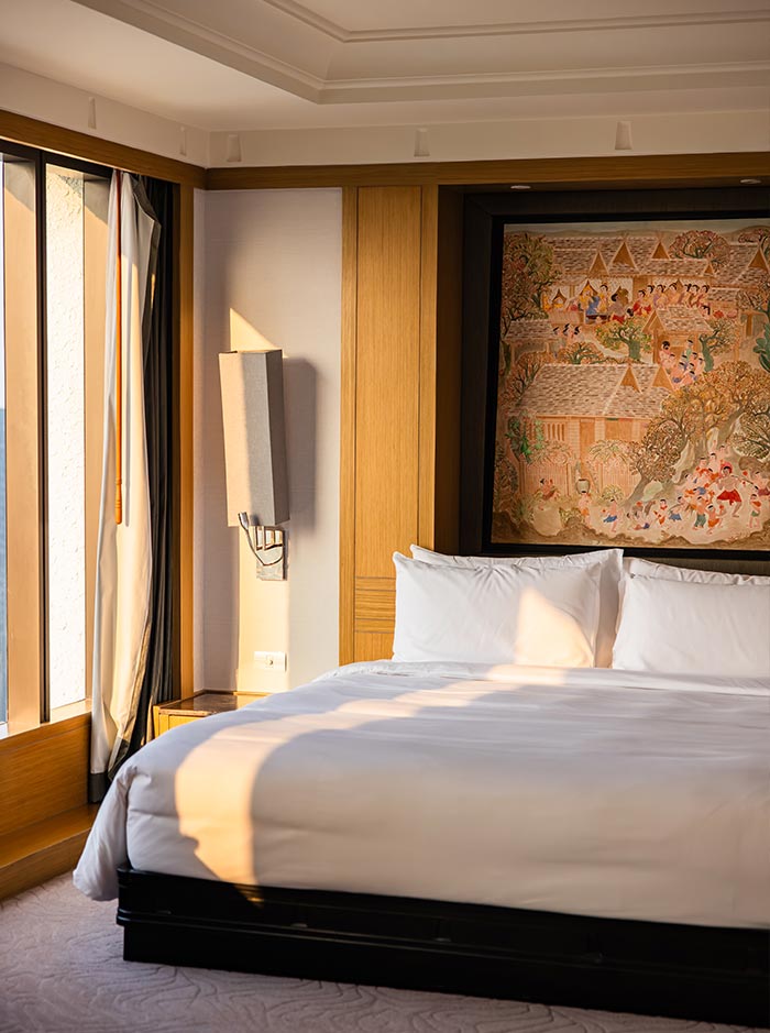 Sunlit hotel bedroom with a neatly made white bed framed by warm wood paneling and soft neutral walls. A large window lets in golden light that falls across the bed, while a framed artwork above the headboard adds a cultural decorative touch. The space feels calm and minimalist, designed for rest and comfort.