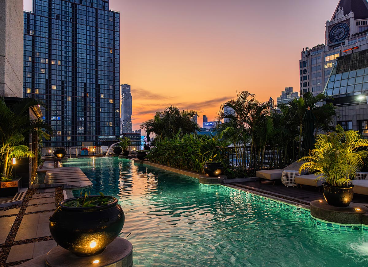 Rooftop infinity pool glowing with soft green lighting at sunset, surrounded by tropical plants and modern lounge seating. Tall city buildings frame the scene, with warm orange and pink tones in the sky reflecting on the water. The atmosphere feels calm and upscale, blending city views with a serene evening retreat.