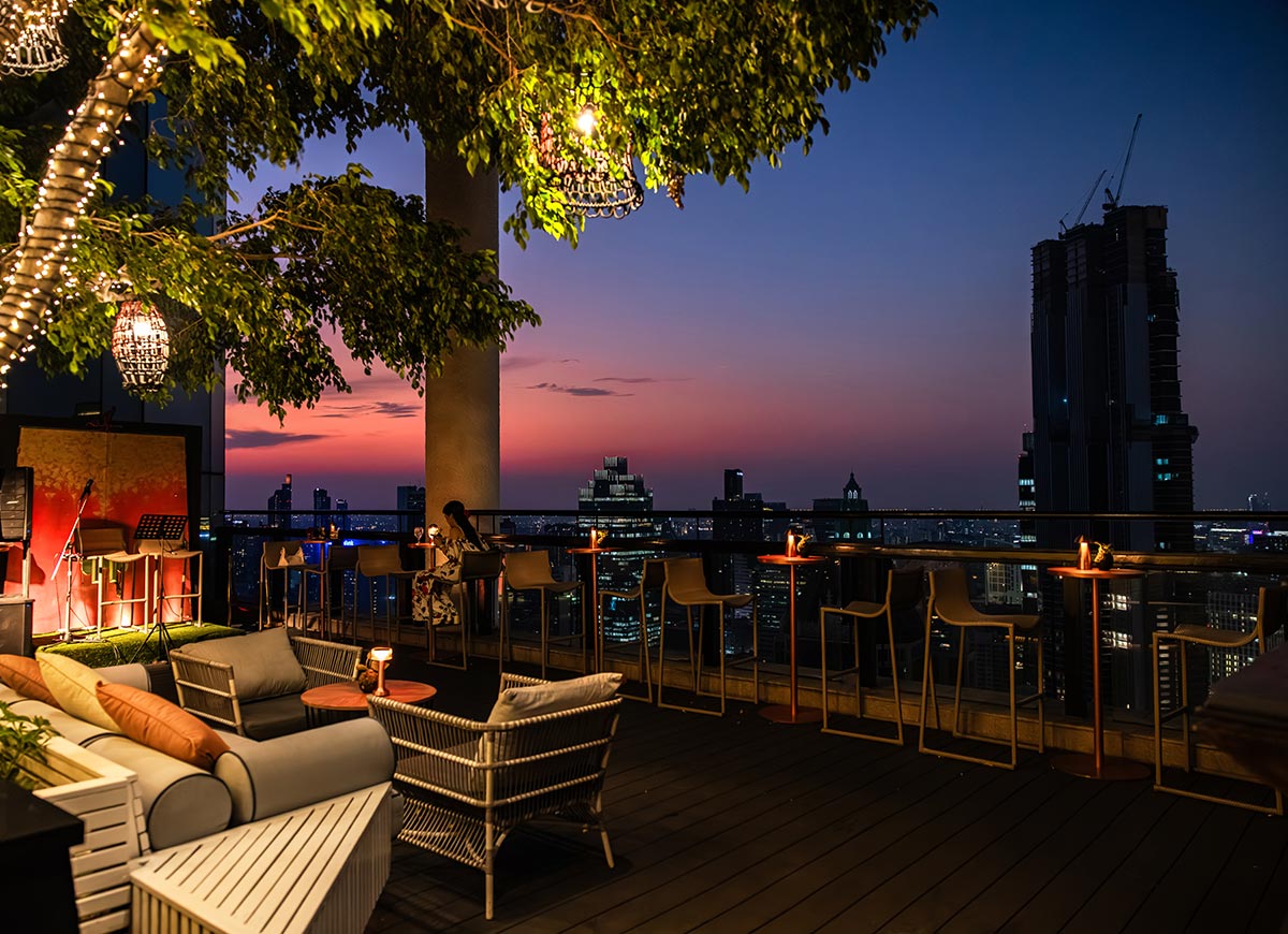 Rooftop bar at sunset with cozy lounge seating and high stools lined along a railing overlooking a city skyline. Warm string lights hang from leafy trees above, while small candles glow on tables creating an intimate evening atmosphere. The sky fades from orange to deep blue as city buildings begin to light up in the background.