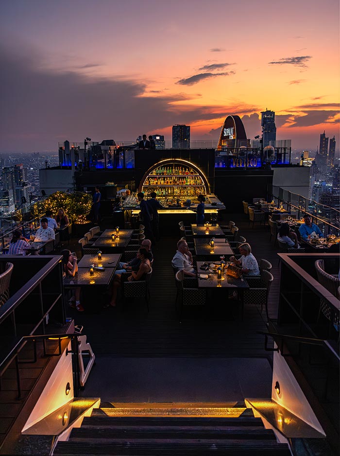 Rooftop restaurant at sunset with candlelit tables and guests dining while overlooking a sprawling city skyline. A glowing bar display with bottles sits at the center beneath a sign reading “VERTIGO,” creating a focal point against the warm orange and purple sky. Steps in the foreground lead up to the lively dining area, adding depth to the elevated evening setting.