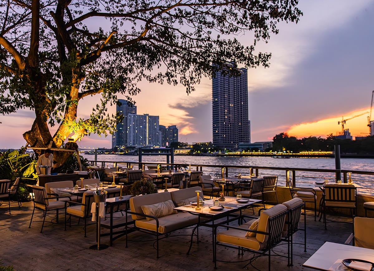 Outdoor riverside restaurant at sunset with neatly set tables and cushioned seating under a large tree wrapped in warm lights. A city skyline rises across the water, reflecting soft orange and purple hues from the evening sky. The atmosphere feels intimate and relaxed, with candlelit tables overlooking the river.