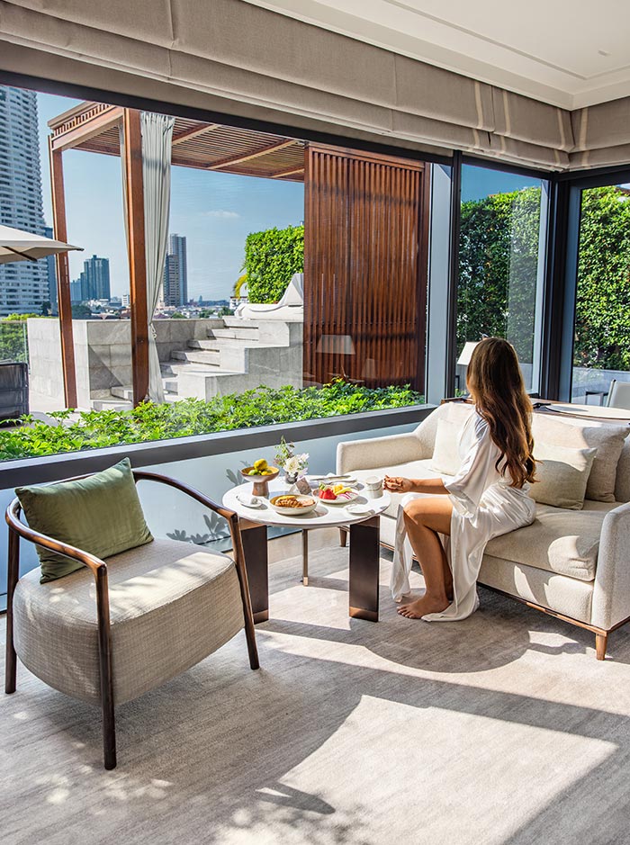 Bright hotel lounge area with a woman in a white robe sitting on a sofa beside a small table set with breakfast. Floor to ceiling windows reveal a private terrace with greenery and a city skyline beyond. Sunlight fills the space, creating a calm and luxurious in room dining atmosphere.