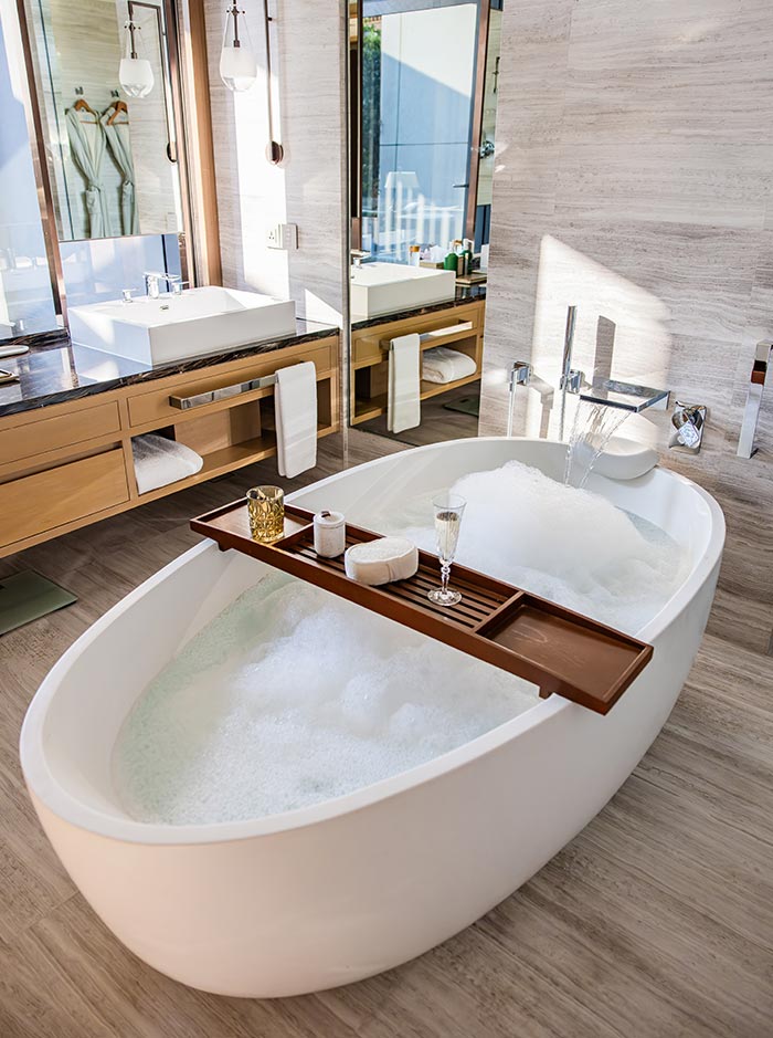 Modern bathroom with a freestanding bathtub filled with bubbles, featuring a wooden tray holding a candle, soap, and a glass. A double vanity with square sinks, mirrors, and neatly folded towels sits in the background against light stone walls. Natural light brightens the clean, spa-like space designed for relaxation.