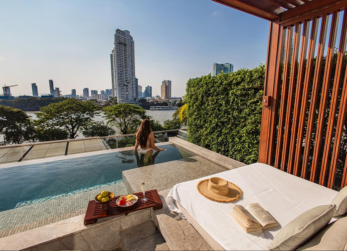 Private plunge pool overlooking a river and city skyline, with a woman sitting at the edge facing the view. A cushioned lounge chair beside the pool holds a straw hat and open book, with a tray of fresh fruit and a drink nearby. The setting feels serene and luxurious, blending a personal retreat with expansive urban scenery.