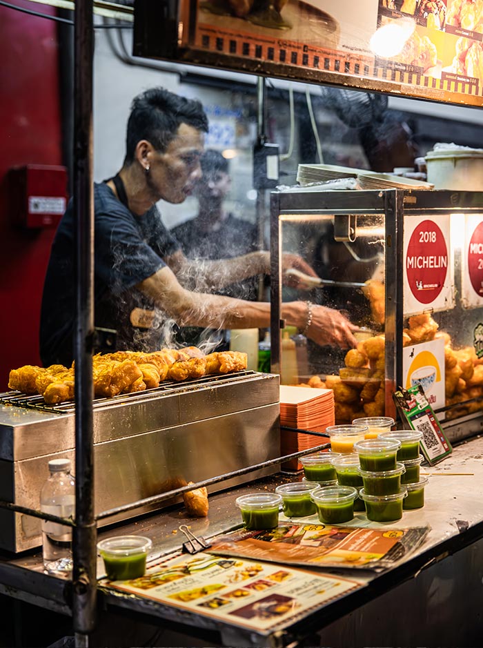Street food vendor frying crispy snacks behind a glass stall at a busy night market. Small cups of green dipping sauce line the counter beside menus and signs including a red “2018 Michelin” sticker displayed on the food cart.