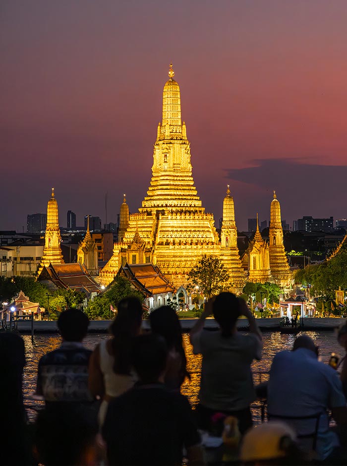 Illuminated temple with towering golden spires glows beside the river at dusk under a purple and pink sky. Silhouettes of people in the foreground watch and photograph the brightly lit temple across the water.