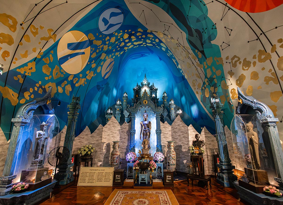 Ornate shrine interior with a central standing statue framed by an elaborate blue and gold arch, surrounded by smaller statues in glass cases. The curved ceiling is painted with celestial patterns and constellations, creating a vivid, immersive backdrop above the detailed altar.