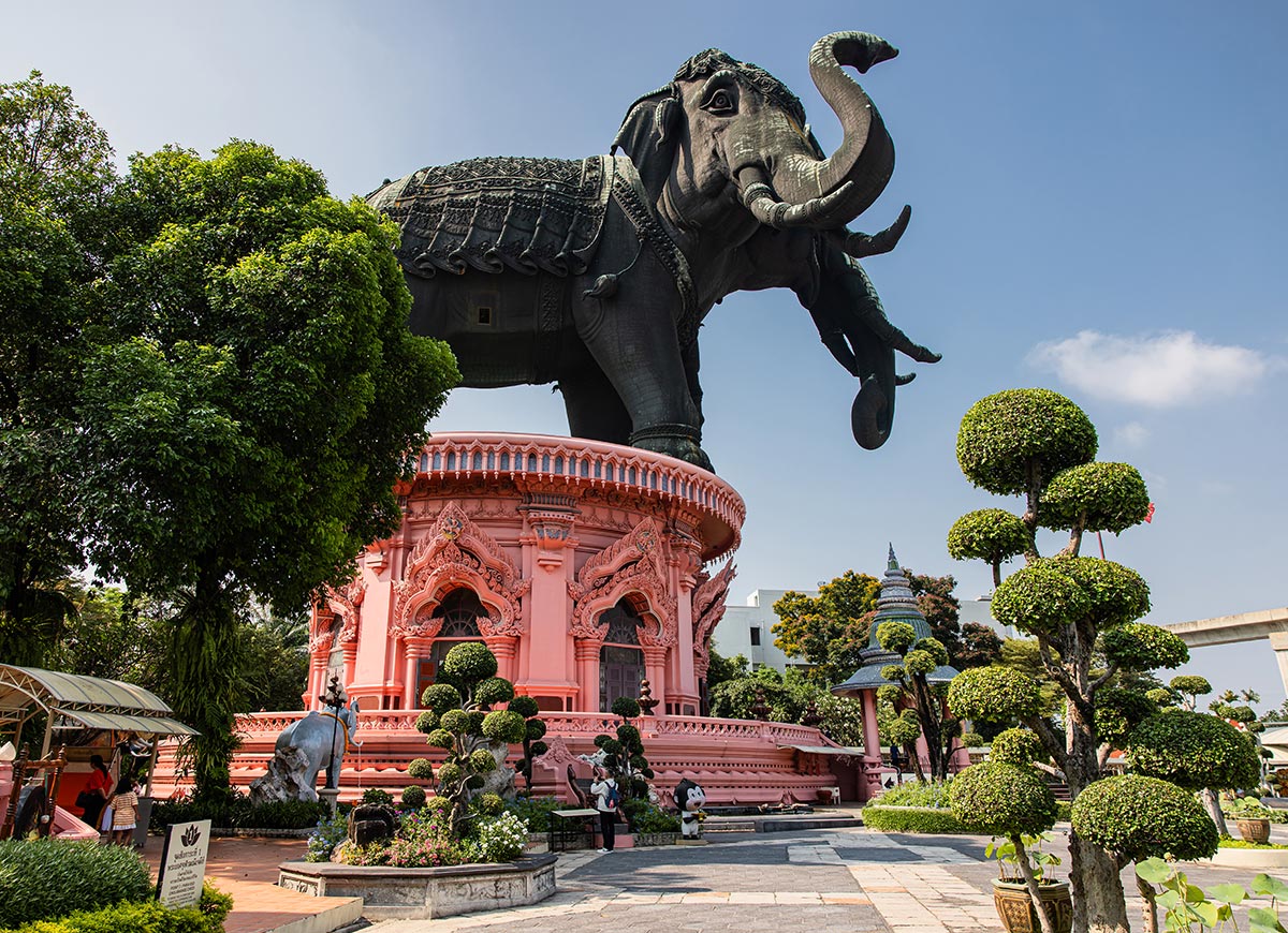 Massive black elephant statue stands atop an ornate pink pedestal building surrounded by manicured trees and garden paths. Visitors walk around the base while the detailed structure rises prominently against a clear blue sky.