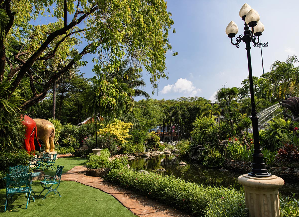 Lush garden with a winding pond surrounded by tropical plants and trees, featuring decorative elephant statues and teal metal benches along a curved pathway. A tall black lamppost stands beside the water as sunlight filters through the greenery on a clear day.