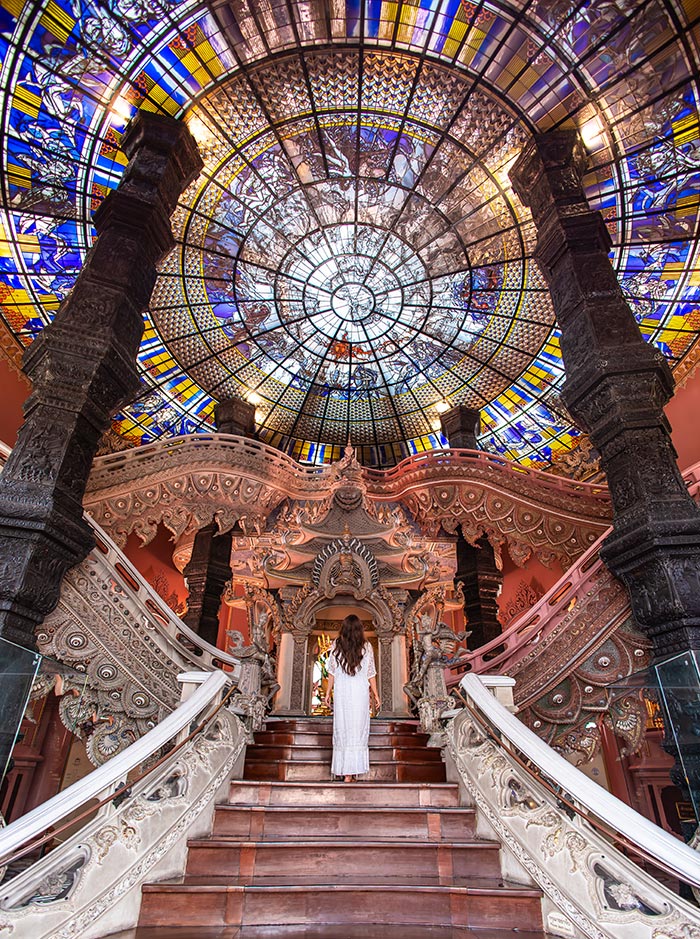 Grand staircase with ornate carvings leads up to a temple interior beneath a large circular stained glass dome filled with intricate blue, gold, and white patterns. A woman in a white dress stands at the top of the steps, centered beneath the detailed ceiling.