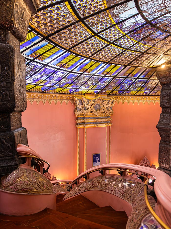 Curved staircase with ornate gold detailing winds beneath a colorful stained glass dome patterned in blue, yellow, and white. Soft light filters through the glass ceiling, illuminating the pink walls and intricate architectural carvings.