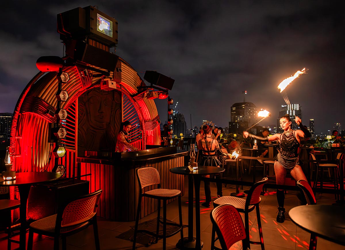 Nighttime rooftop bar with red ambient lighting and a DJ booth framed by curved industrial decor. A fire dancer performs with flaming torches in front of guests while the Bangkok skyline glows in the background.