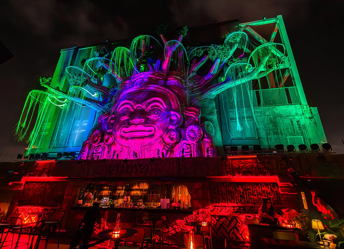 Massive neon lit sculpture with a glowing pink face and tree like branches towers above an outdoor rooftop bar at night. Red and green lighting illuminates the futuristic venue while guests sit beneath the dramatic installation surrounded by dark city skies.