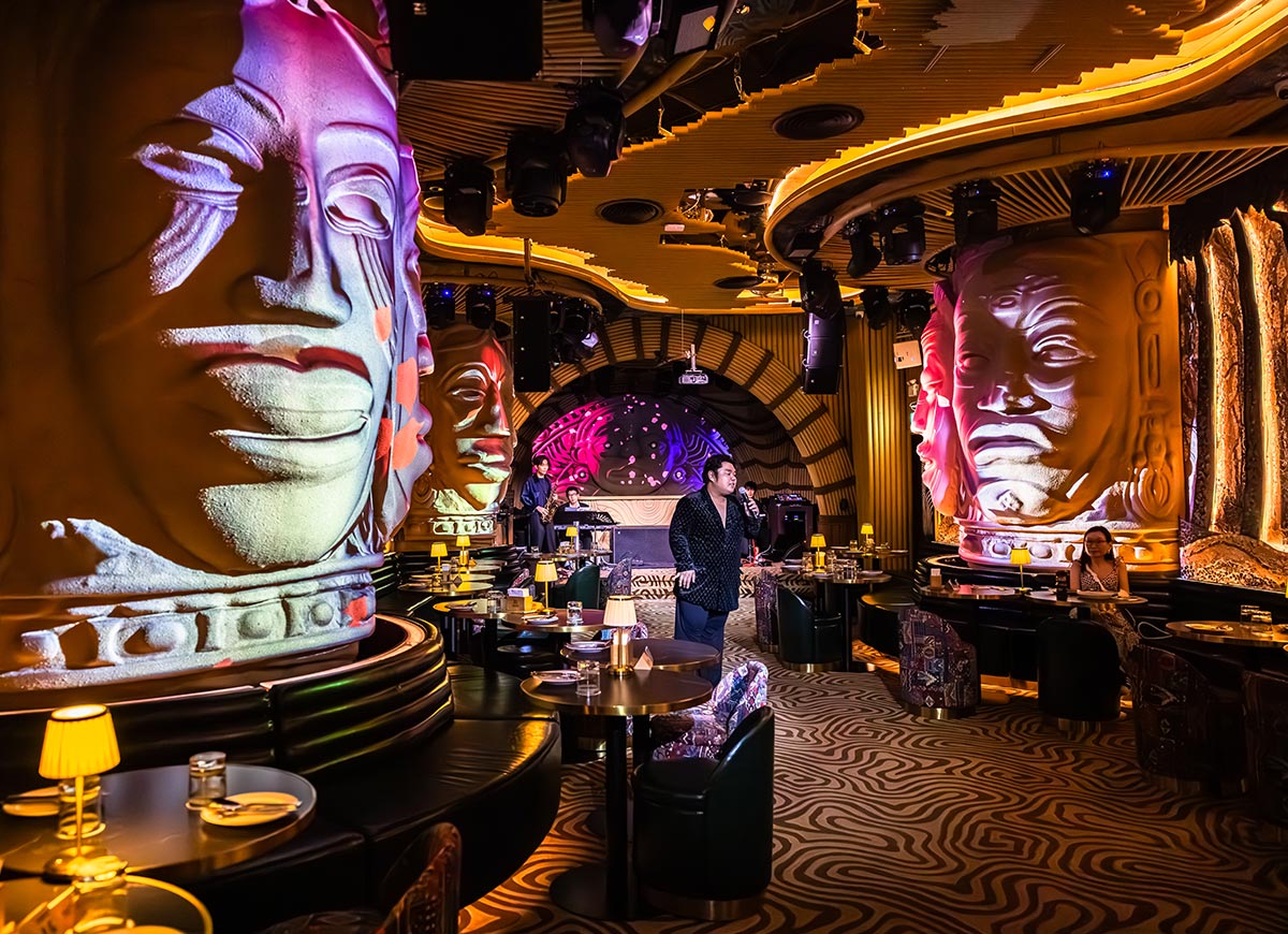 Dramatic restaurant interior with towering sculptural faces illuminated in pink and white lighting surrounding an intimate dining space. A live singer performs near the stage while curved golden ceiling details and patterned floors create a futuristic nightlife atmosphere.