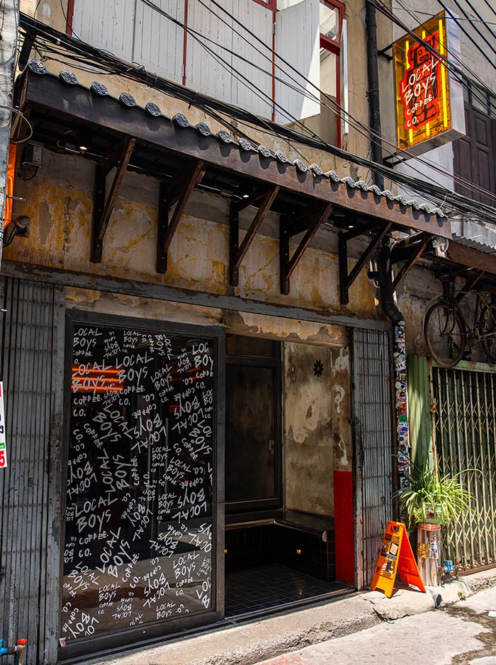 Street-front café with a weathered facade and a black door covered in repeated white text reading “LOCAL BOYS.” A neon sign above reads “LOCAL BOYS COFFEE CO.” while an orange sidewalk sign stands near the entrance.