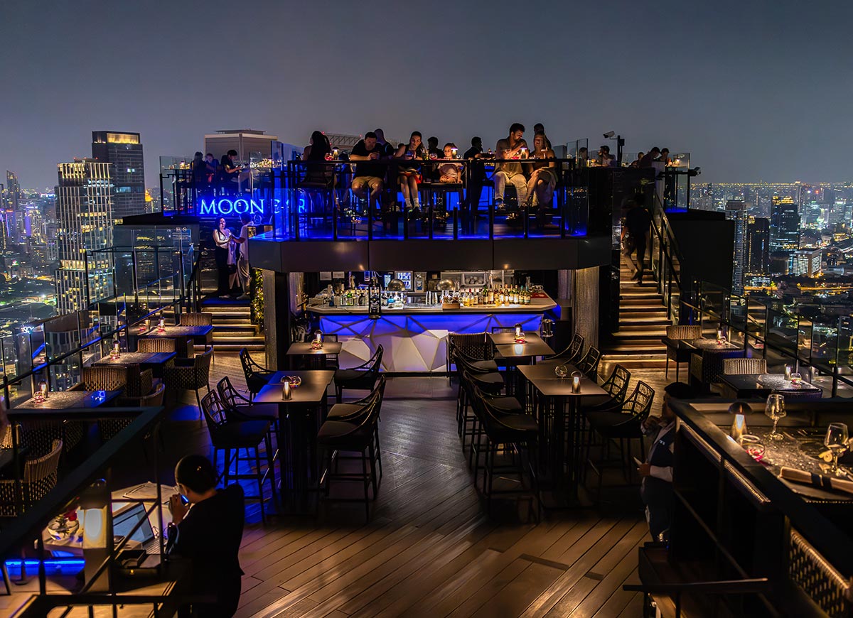 Modern rooftop bar with glowing blue lighting and multiple terrace levels overlooking a brightly lit city skyline at night. Guests sit at tables and gather around the central bar while neon signs illuminate the upscale open air venue.