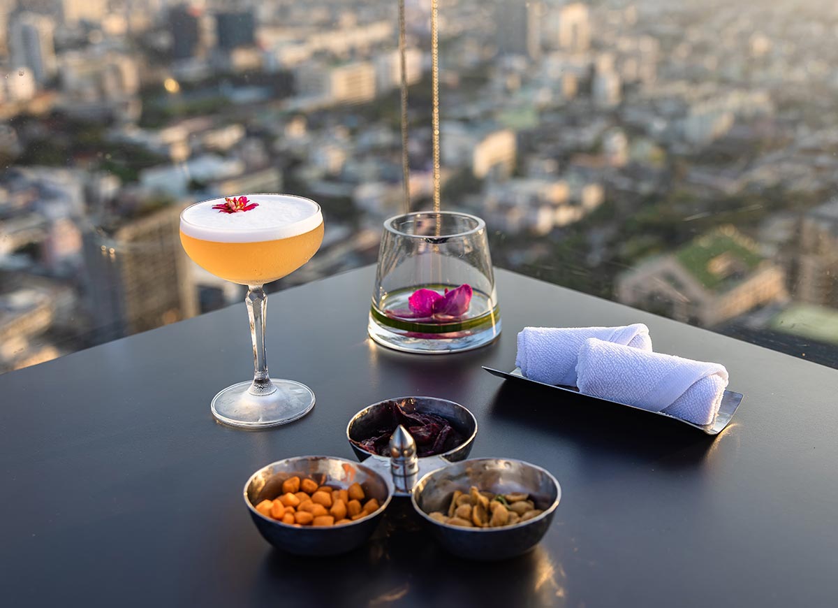 Elegant cocktail with a foamy top and edible flower garnish sits on a rooftop table overlooking a blurred city skyline. Small bowls of nuts and dried fruit, rolled hand towels, and a glass with floating flower petals complete the upscale dining setup.