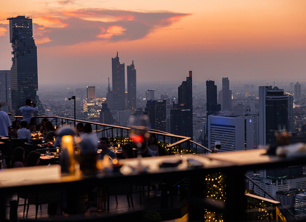 Rooftop bar with candlelit tables overlooks a dense city skyline at sunset, with warm orange and pink hues fading into evening. Guests sit and mingle along the terrace while tall buildings rise against the glowing sky.