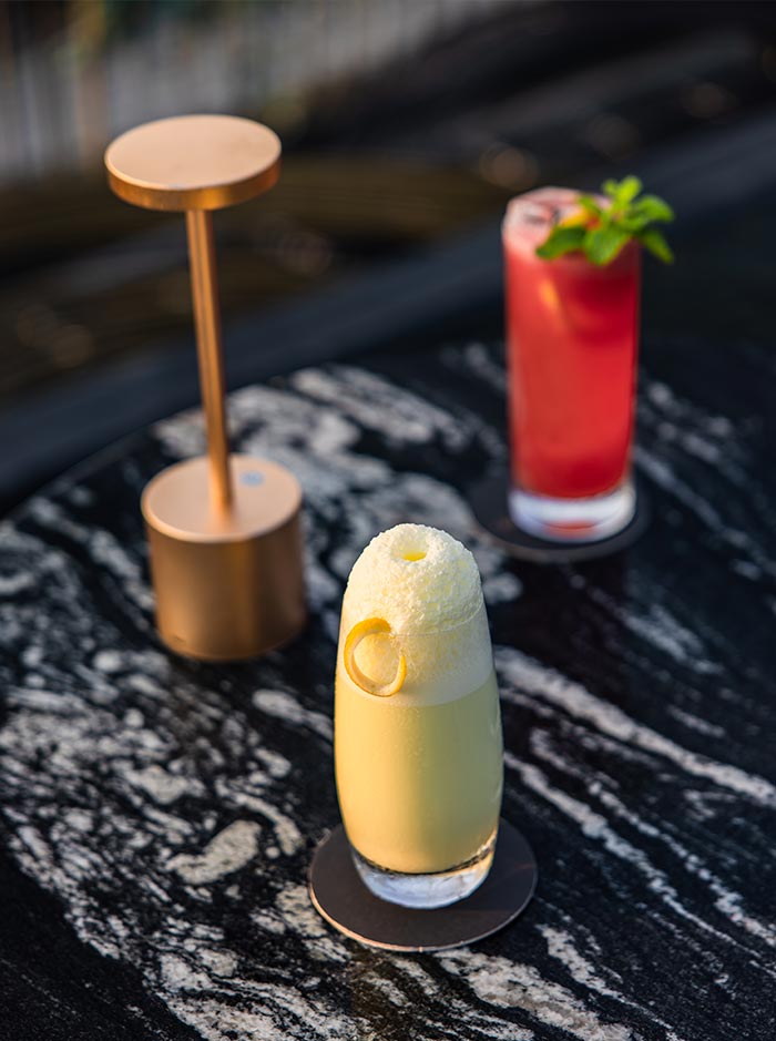 Two colorful cocktails served on a black marble table at a rooftop bar with soft evening lighting. A creamy yellow drink topped with foam and citrus garnish sits in the foreground while a bright pink cocktail with mint leaves is blurred in the background beside a modern gold lamp.