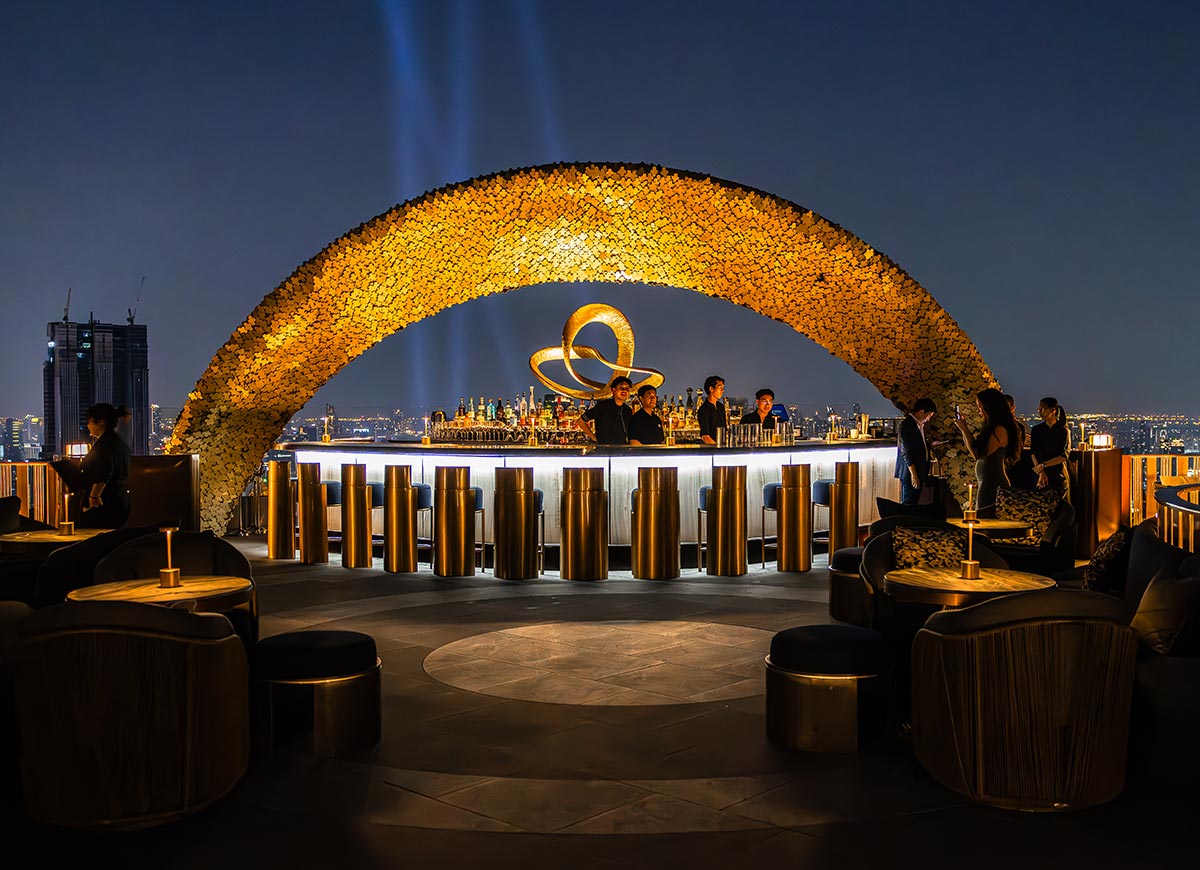 Elegant rooftop cocktail bar illuminated at night with a glowing golden arch framing the circular bar area. Guests sit in modern lounge chairs beneath warm ambient lighting while bartenders serve drinks against the sparkling Bangkok skyline.