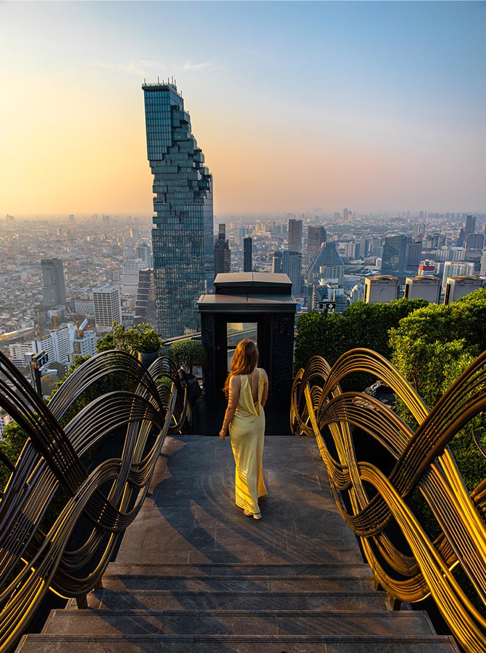 Woman in a flowing yellow dress walks down a staircase with sculptural gold railings toward a rooftop viewpoint overlooking a sprawling city skyline. A tall modern skyscraper rises prominently against a soft sunset sky in the background.