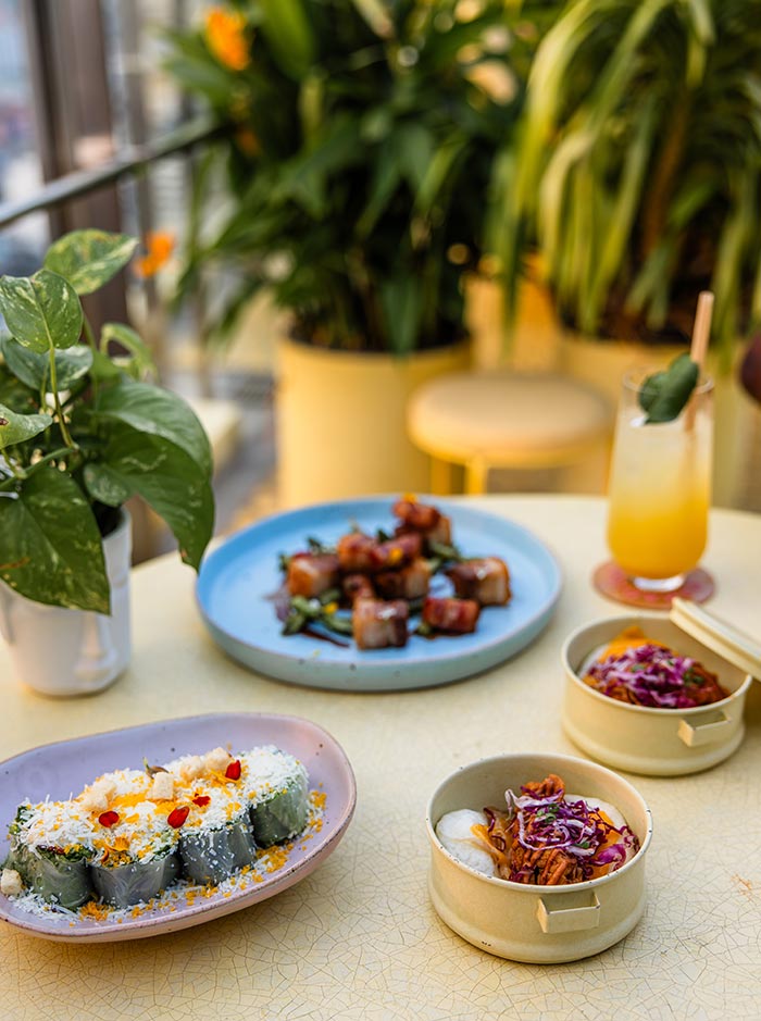 Colorful spread of modern fusion dishes and cocktails served on a table surrounded by lush indoor plants. Plates of sushi rolls, tacos, and glazed bites are arranged beside tropical drinks in a bright restaurant with a relaxed garden inspired atmosphere.