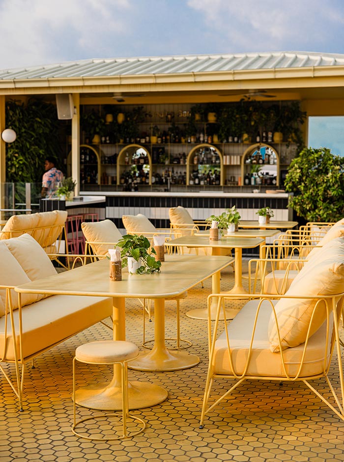 Sunny rooftop bar with cream colored lounge seating and yellow metal chairs arranged around modern tables. Warm golden light illuminates the open air terrace while the bar lined with plants and bottles creates a relaxed tropical atmosphere.