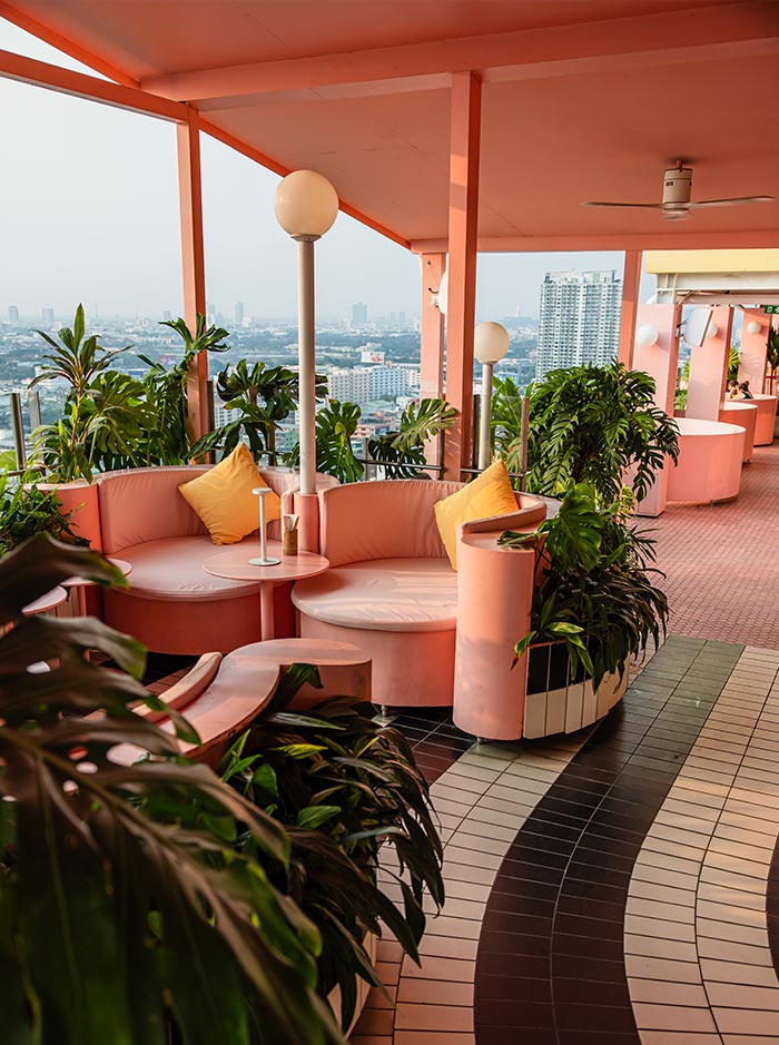 Pastel pink rooftop lounge with curved booth seating, tropical plants, and panoramic city views in Bangkok. Soft sunset light warms the retro inspired space while black and white tiled flooring leads through the open air terrace.
