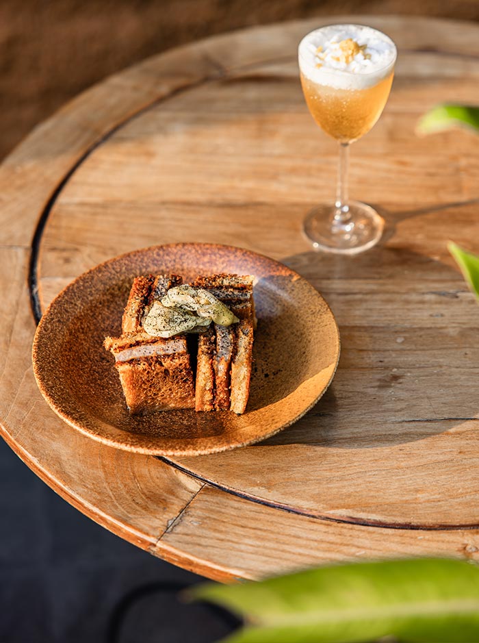Rustic plated dessert made with layered toasted bread and cream served on a wooden table beside a sparkling golden cocktail topped with foam and flower petals. Warm sunset lighting highlights the textured pastry and elegant rooftop drink presentation.