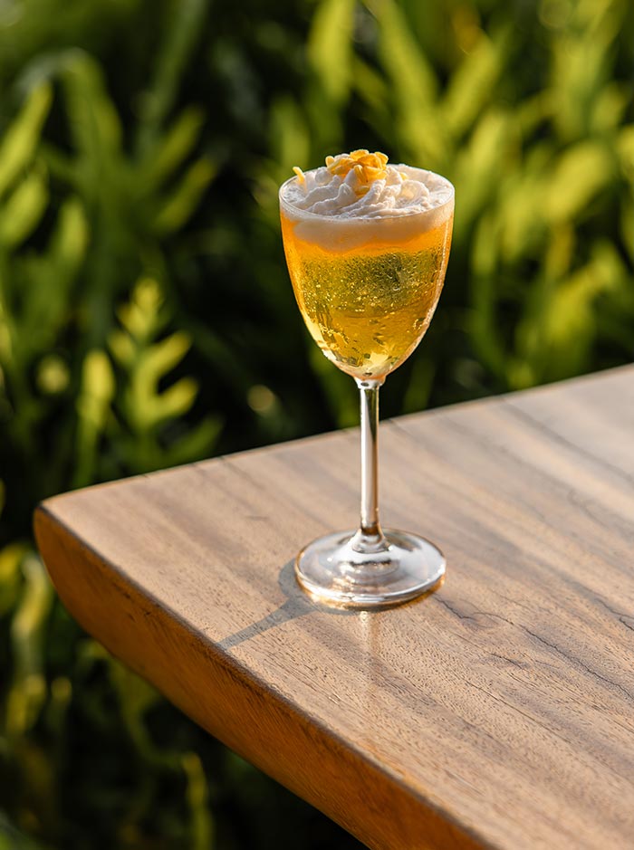 Golden sparkling cocktail topped with whipped foam and yellow flower petals served in a wine glass on a wooden rooftop table. Soft evening sunlight highlights the bubbly drink while blurred tropical greenery creates a relaxed outdoor atmosphere.