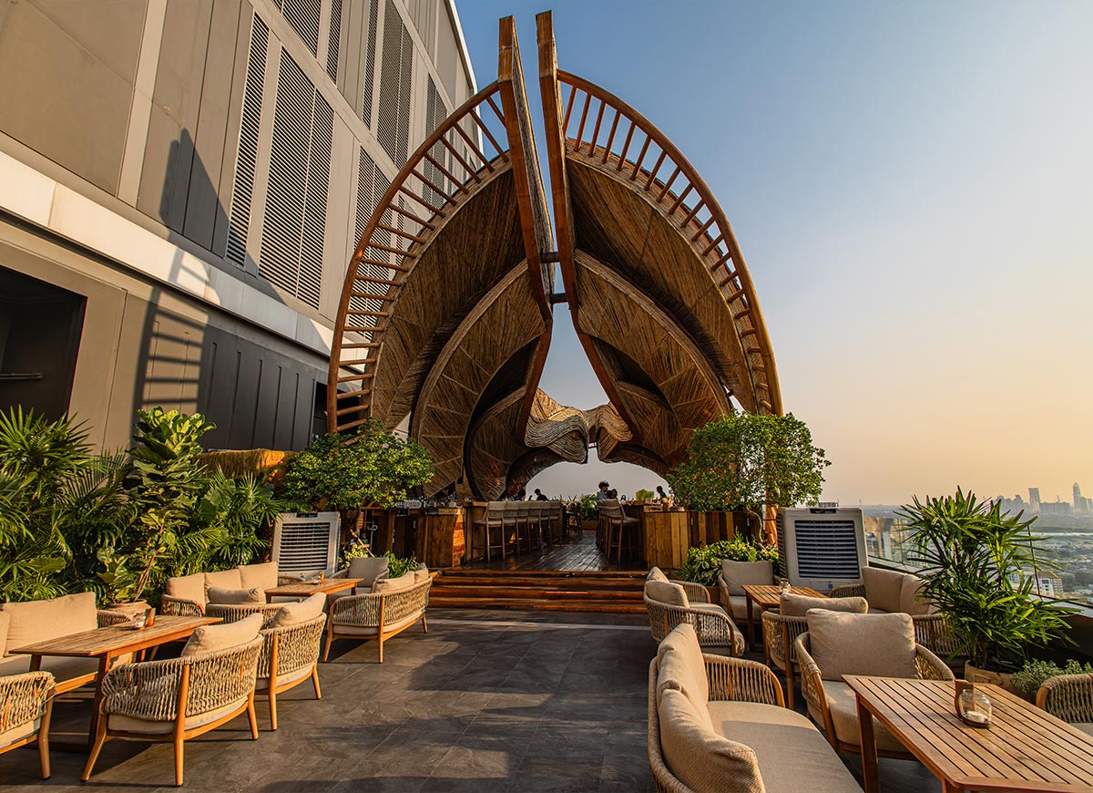 Open air rooftop lounge with curved woven wood architecture rising above cozy seating areas and tropical plants in Bangkok. Warm golden sunlight illuminates the sculptural canopy while guests relax beneath the dramatic organic design overlooking the city skyline.