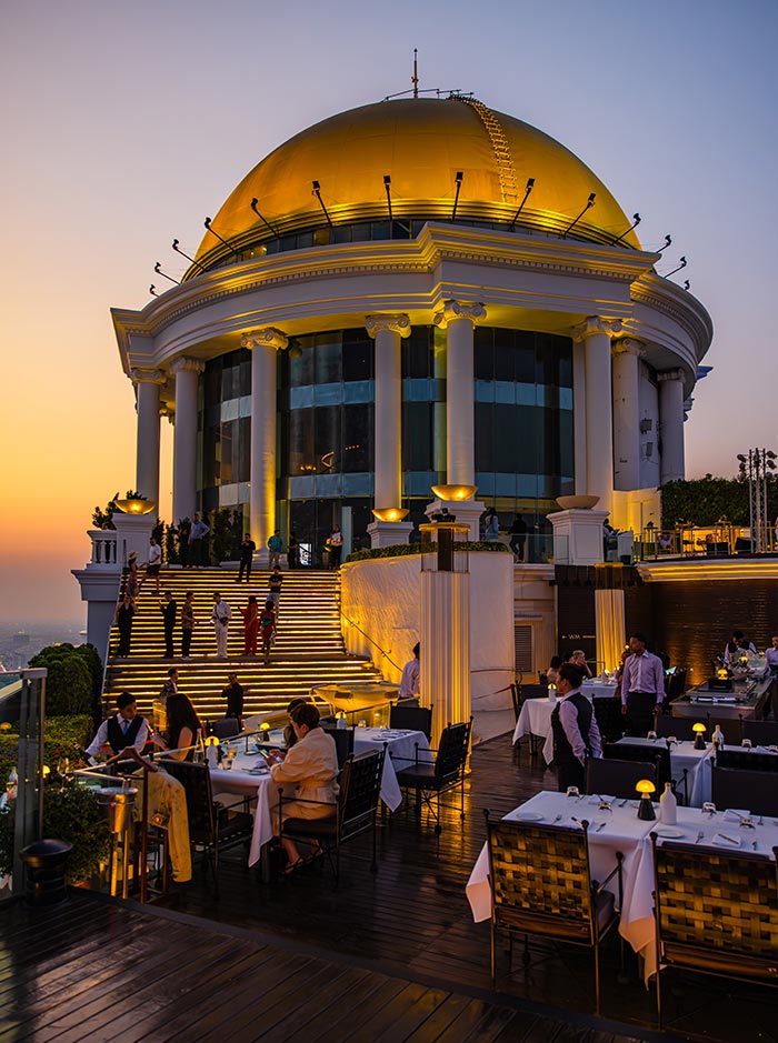 Luxury rooftop restaurant with white tablecloth tables and glowing staircase leading to a grand domed building illuminated in gold at sunset. Diners and staff gather on the terrace while the soft evening sky fades into warm pastel tones above the city.