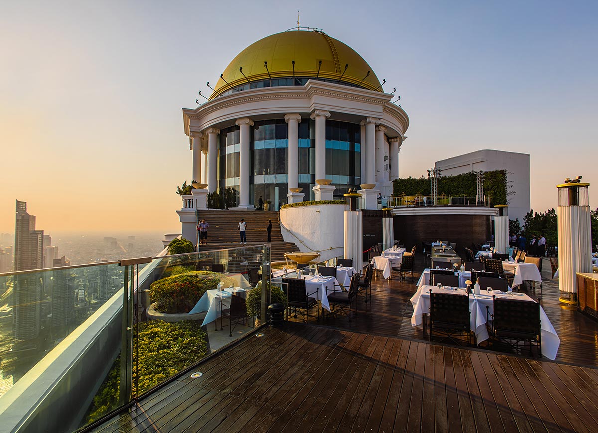 Elegant rooftop restaurant terrace with white tablecloth dining tables overlooks the city skyline at sunset. A grand domed building with tall white columns rises above illuminated steps, creating a luxurious atmosphere against the soft evening sky.