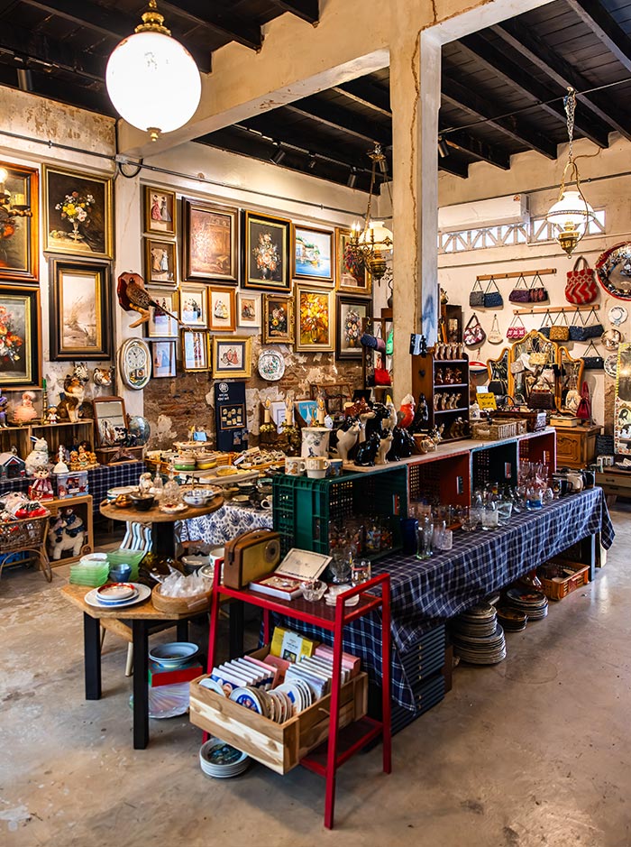 Cozy vintage shop interior filled with shelves and tables displaying ceramics, glassware, framed artwork, and collectibles. Walls are covered with assorted paintings while hanging lights illuminate the eclectic mix of antiques and decorative items.