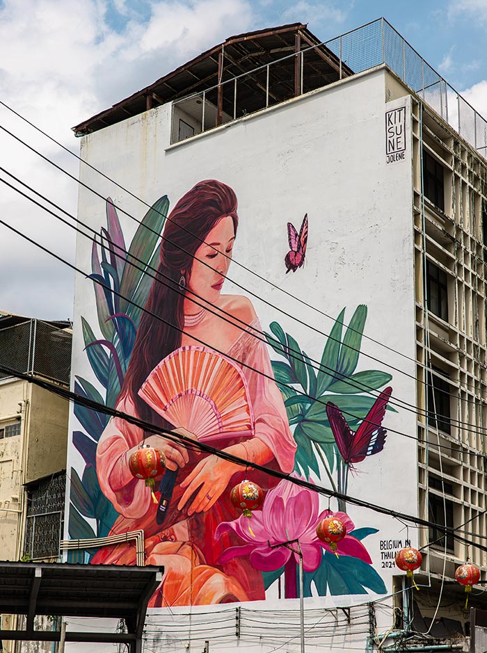 Large mural of a woman in a pink outfit holding a fan is painted on the side of a white building, surrounded by tropical leaves and butterflies. Overhead wires and hanging red lanterns cross in front of the artwork, with a small sign reading “KITSUNE JOLENE” near the top.