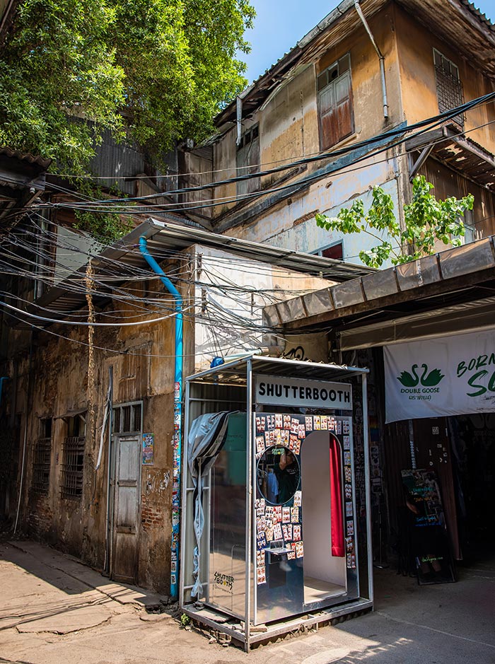 Weathered alleyway with tangled overhead wires and aged buildings frames a small glass booth labeled “SHUTTERBOOTH” covered in photo strips. Sunlight highlights the narrow passage and textured walls, contrasting with the modern photo booth setup.