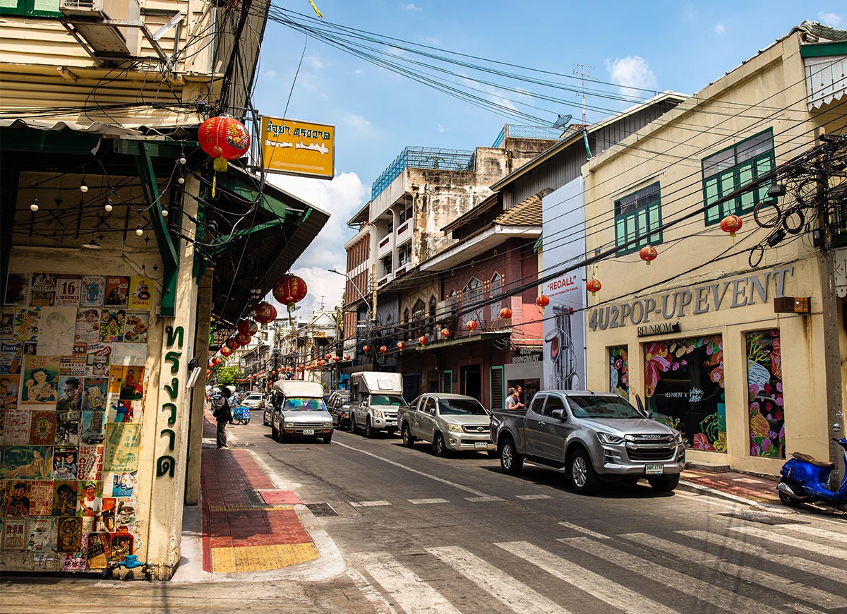 Busy street lined with low-rise buildings decorated with red hanging lanterns strung across the road. Cars drive past small shops including one with a sign reading “4U2POP-UP EVENT” while overhead wires crisscross against a blue sky.