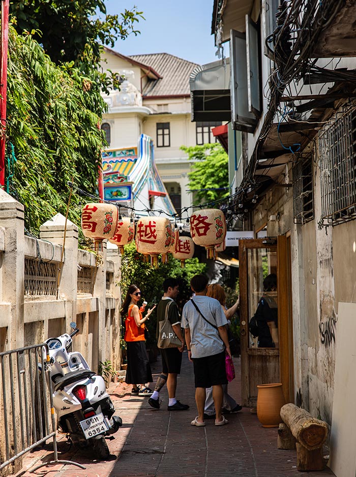 Narrow alleyway with hanging lanterns displaying red Chinese characters stretches between weathered buildings and greenery. A small group of people stands near a doorway while a parked scooter lines the brick path under bright daylight.