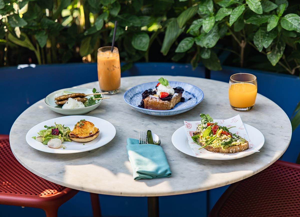 Round marble table set with brunch dishes including avocado toast, eggs benedict, skewers, and a dessert topped with berries and cream. Iced coffee and orange juice sit alongside the plates, with a folded napkin and cutlery in the center. Lush green plants surround the cozy outdoor seating area, creating a relaxed garden dining atmosphere.