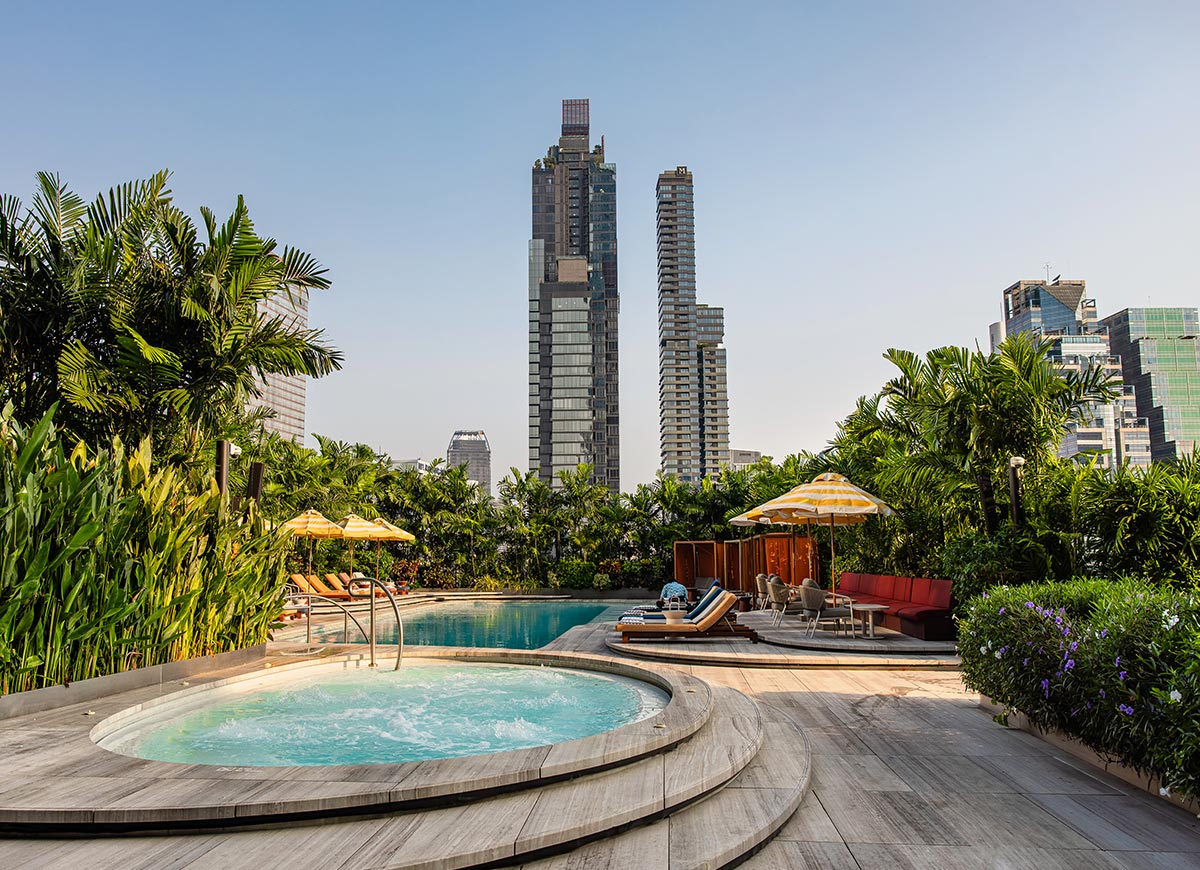 Rooftop pool with a circular hot tub in the foreground, surrounded by wooden decking, lounge chairs, and yellow striped umbrellas. Lush tropical plants line the space, with modern high rise buildings rising in the background under a clear sky. The setting feels like a quiet urban oasis designed for relaxation above the city.
