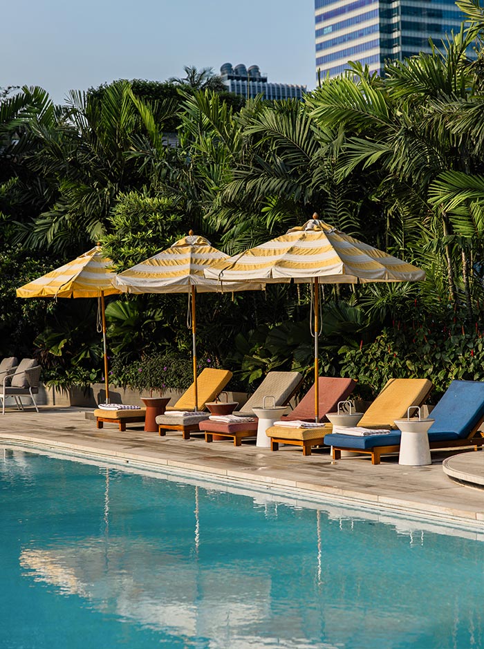 Poolside lounge chairs in warm tones line the edge of a clear blue swimming pool, shaded by yellow striped umbrellas. Dense tropical greenery forms a lush backdrop, with a glimpse of city buildings rising above the trees. The setting blends resort style relaxation with an urban rooftop environment.