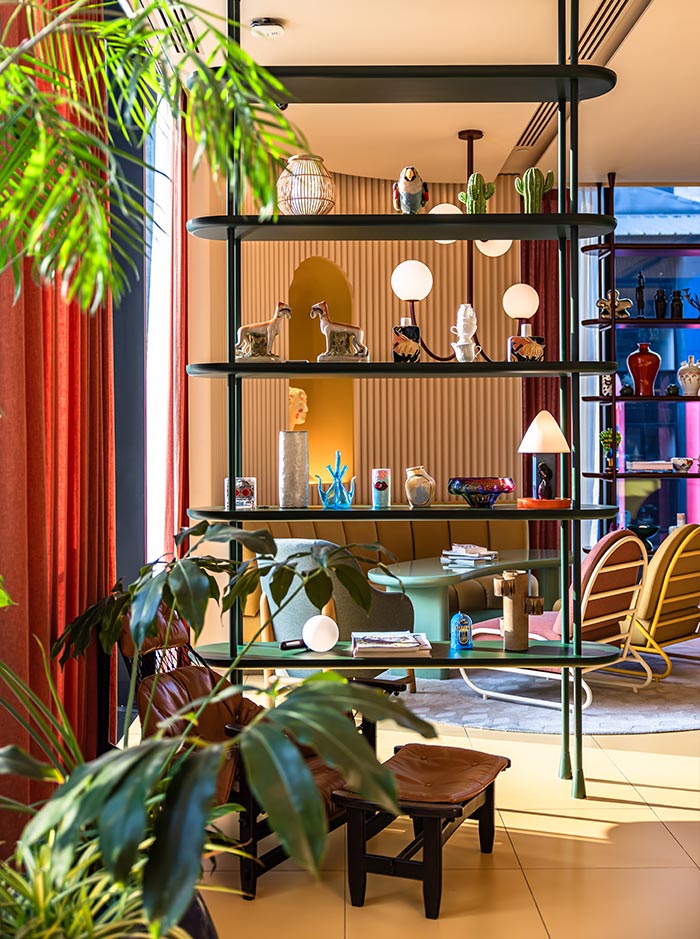 Open shelving unit styled with eclectic decor including small sculptures, vases, and lamps, set within a colorful modern living space. Warm tones, curved architectural details, and layered textures create a vibrant and artistic interior. Large leafy plants in the foreground partially frame the scene, adding a lush and relaxed feel.