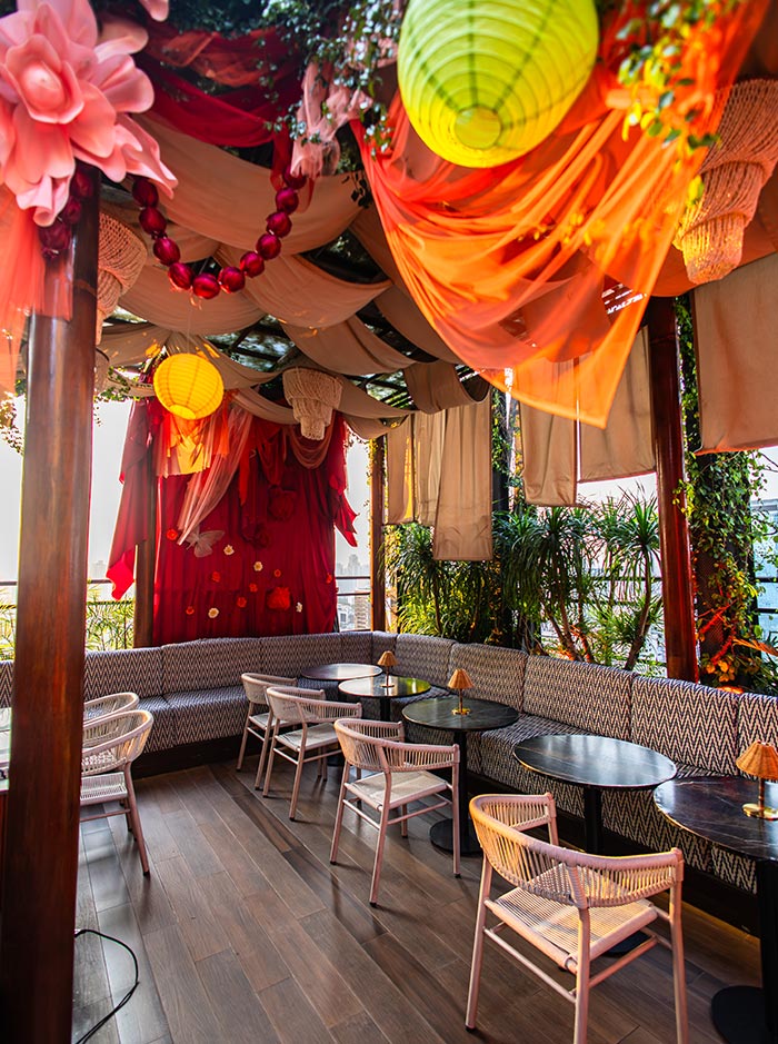 Cozy rooftop lounge area with pink chairs and black tables sits beneath flowing orange and cream fabric draped across the ceiling. Colorful lanterns, oversized floral decorations, and hanging greenery create a vibrant and whimsical atmosphere filled with warm sunlight.
