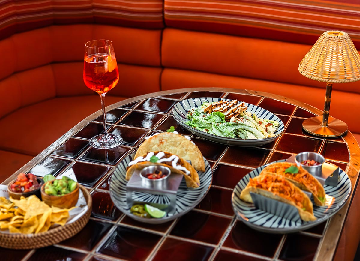 Restaurant table with glossy dark tile surface displays tacos, salad, chips with guacamole and salsa, and a red cocktail in a wine glass. Warm orange booth seating and a woven table lamp create a cozy, upscale dining atmosphere.
