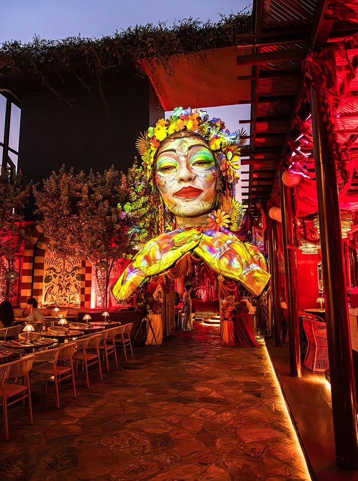 Large colorful sculpture of a serene woman’s face with floral details hangs above a stylish rooftop restaurant illuminated with warm red lighting. Dining tables line the open air space while dramatic evening lighting creates a vibrant and artistic atmosphere.
