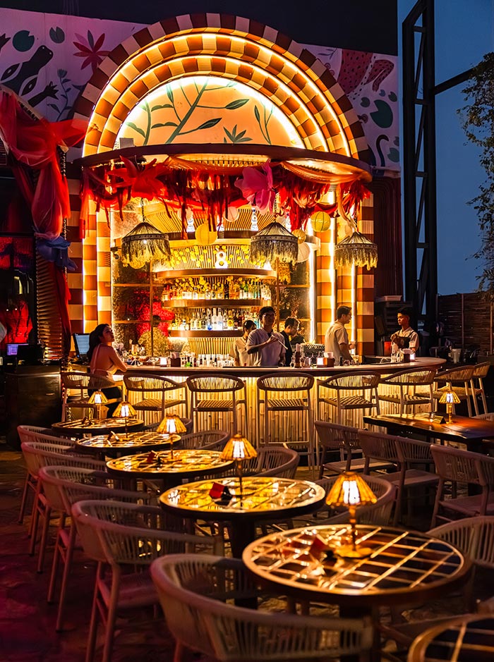 Glowing rooftop bar with warm golden lighting features an ornate illuminated counter framed by colorful arch details and hanging decorations. Candlelit tables and woven chairs fill the stylish open air dining space as guests gather around the vibrant bar at night.
