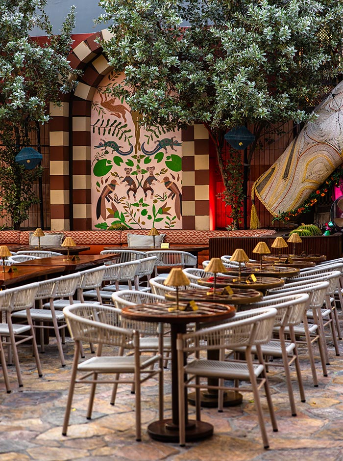 Outdoor restaurant patio with woven white chairs and round wooden tables arranged beneath leafy trees and warm accent lighting. A decorative mural featuring monkeys and tropical plants adds a playful artistic backdrop to the stylish open air dining space.